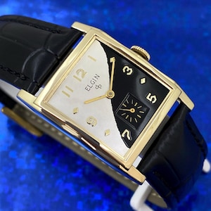 Peut inclure: Une montre Elgin vintage avec un bo&icirc;tier rectangulaire dor&eacute; et un bracelet en cuir noir. Le cadran est divis&eacute;, avec un c&ocirc;t&eacute; blanc avec des chiffres dor&eacute;s et le logo Elgin, et un c&ocirc;t&eacute; noir avec des chiffres dor&eacute;s et un petit cadran de secondes.