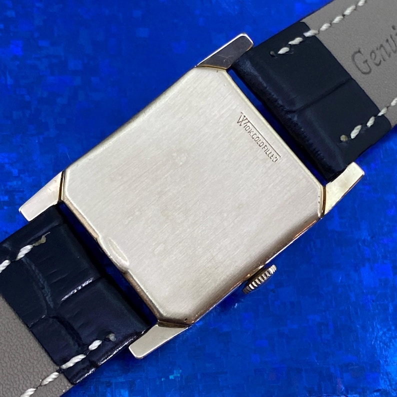 Peut inclure: Gros plan d'une montre vintage dor&eacute;e avec un cadran rectangulaire. Le dos de la montre est estampill&eacute; "10K GOLD FILLED" et est fix&eacute; &agrave; un bracelet en cuir noir avec des coutures blanches. La montre est sur fond bleu.