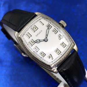 Pu&ograve; includere: Un orologio da polso Gruen in argento con quadrante bianco e cinturino in pelle nera. L'orologio ha indici delle ore in numeri romani e indici dei minuti in numeri arabi.