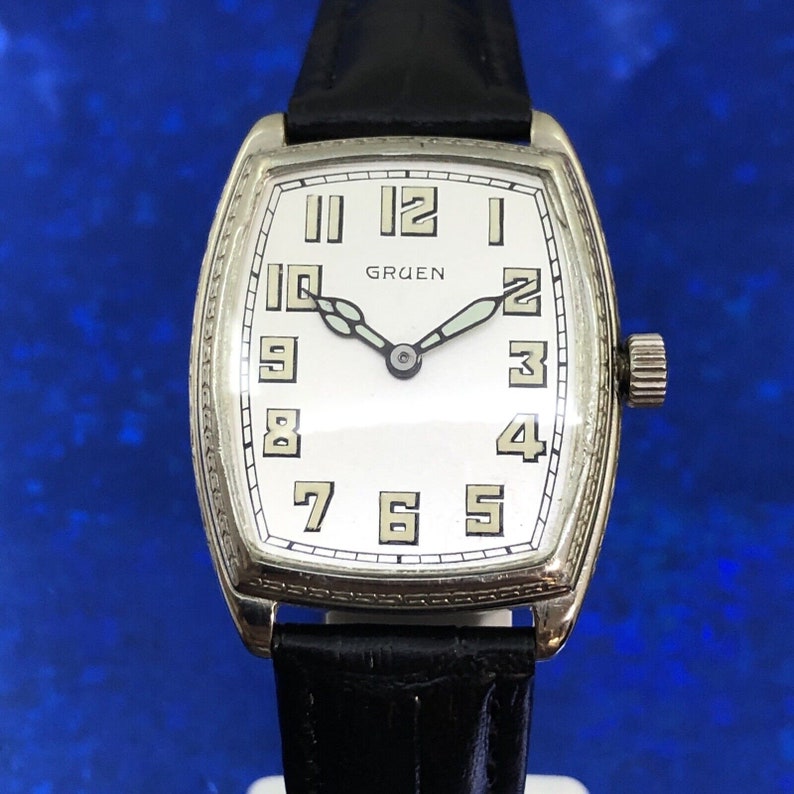 Pu&ograve; includere: Un orologio da polso Gruen argentato con quadrante bianco e lancette nere. L'orologio ha una cassa rettangolare e un cinturino in pelle nera.