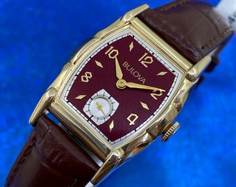 Könnte beinhalten: Eine Vintage-Bulova-Armbanduhr mit einem rechteckigen goldfarbenen Gehäuse und einem weinroten Zifferblatt. Das Zifferblatt hat goldene Zeiger und Ziffern sowie ein kleines Hilfszifferblatt für die Sekunden. Die Uhr hat ein braunes Lederarmband.