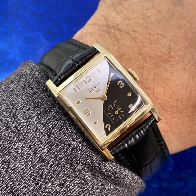 Peut inclure: Une montre Elgin vintage avec un bo&icirc;tier rectangulaire dor&eacute; et un bracelet en cuir noir. Le cadran de la montre est divis&eacute; en diagonale, avec un design argent&eacute; et noir. Le c&ocirc;t&eacute; argent&eacute; porte les chiffres 1, 3, 7, 9, 11 et 12.