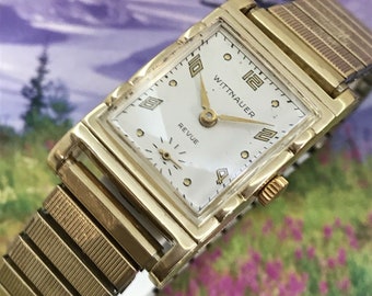 May include: A gold-toned Wittnauer Revue wristwatch with a square face and a gold-toned metal band. The watch face is white with black Roman numeral hour markers and black hands.