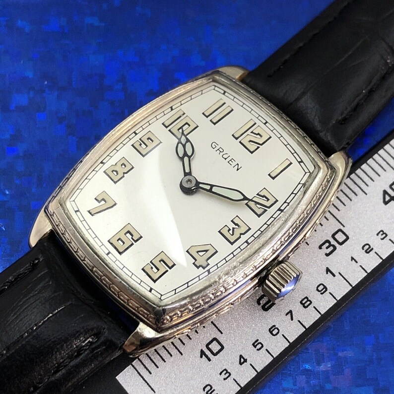 Pu&ograve; includere: Un orologio da polso Gruen argentato con quadrante bianco e lancette nere. L'orologio ha una cassa rettangolare e un cinturino in pelle nera. Il quadrante dell'orologio ha indici orari in numeri romani e indici dei minuti in numeri arabi.
