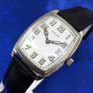 Op de afbeelding: Een zilveren Gruen polshorloge met een witte wijzerplaat en zwarte wijzers. Het horloge heeft een rechthoekige kast en een zwarte leren band.