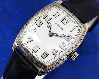 May include: A silver Gruen wristwatch with a white face and black hands. The watch has a rectangular case and a black leather band.