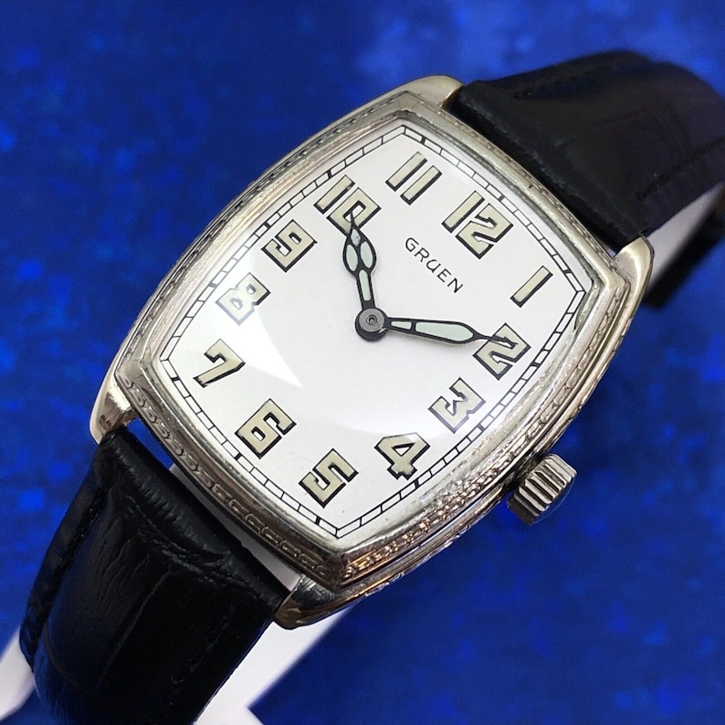 Pu&ograve; includere: Un orologio da polso Gruen argentato con quadrante bianco e lancette nere. L'orologio ha una cassa rettangolare e un cinturino in pelle nera.