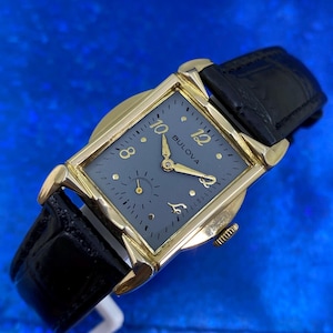 Op de afbeelding: Een vintage Bulova horloge met een rechthoekige goudkleurige kast en een donkergrijze wijzerplaat. Het horloge heeft gouden wijzers en uurmarkeringen, een kleine secondenwijzer en een zwarte leren band. Het woord "Bulova" staat op de wijzerplaat.