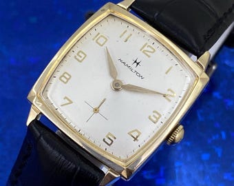 Può includere: Un orologio da polso Hamilton vintage dorato con quadrante quadrato e bianco. L'orologio presenta lancette e numeri dorati, un cinturino in pelle nera e il logo Hamilton. Un orologio classico.