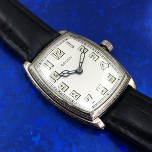 Pu&ograve; includere: Un orologio da polso Gruen argentato con quadrante bianco e lancette nere. L'orologio ha una cassa rettangolare e un cinturino in pelle nera.