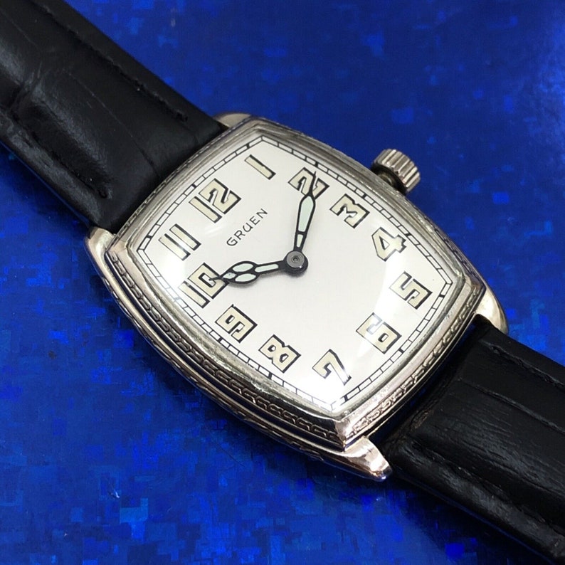Pu&ograve; includere: Un orologio da polso Gruen argentato con quadrante bianco e lancette nere. L'orologio ha una cassa rettangolare e un cinturino in pelle nera.