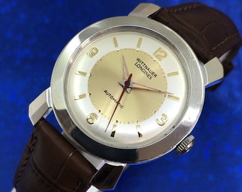 Puede incluir: Un reloj de pulsera plateado con una correa de cuero marrón. La esfera del reloj es blanca con detalles dorados y el texto "Wittnauer Longines Automatic" está impreso en la esfera.