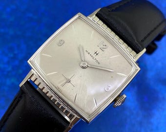 May include: A vintage Hamilton wristwatch with a silver square face and black leather band. The watch face features silver hands, hour markers, and the Hamilton logo. The watch has a silver bezel and a crown on the side.