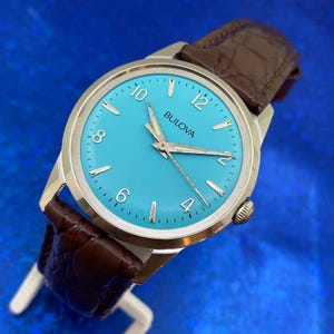 May include: A Bulova wristwatch with a turquoise face, silver case, and brown leather band. The watch face features silver hands and numerals. The watch is a classic design with a textured leather band.