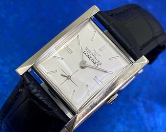 May include: A rectangular silver-tone wristwatch featuring a white dial with silver-tone hour markers and hands. The dial displays the text WITTNAUER LONGINES. The watch includes a black leather strap with a textured finish and a small sub-dial.