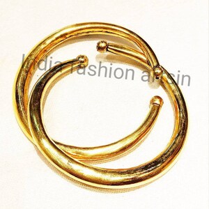 May include: Two gold-colored metal bangles, one slightly larger than the other, are stacked on top of each other. The bangles are smooth and have a rounded shape.