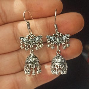 May include: A pair of silver earrings with a butterfly design and a bell-shaped dangle. The earrings have a silver hook closure.