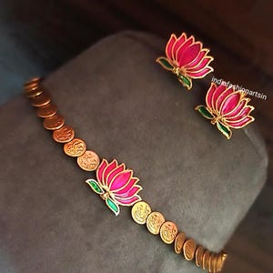 May include: A gold-toned necklace and matching earrings set. The necklace features a series of small, round, coin-like elements and a central lotus flower pendant with pink and green enamel detailing. The earrings are lotus flower studs with similar pink and green enamel.