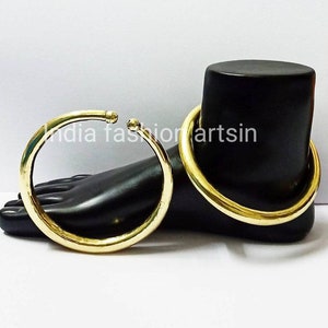 May include: Two gold-colored metal bangles, one larger than the other, on a black mannequin foot. The bangles are smooth and round. The text "india fashionartsin" is visible on the mannequin's foot.
