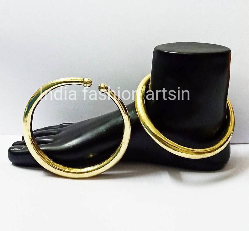 May include: Two gold-colored metal bangles, one larger than the other, on a black mannequin foot. The bangles are smooth and round. The text "india fashionartsin" is visible on the mannequin's foot.