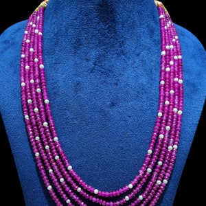 Op de afbeelding: Een meerrijige ketting gemaakt van roze en witte kralen. De ketting is gemaakt van vijf rijen roze kralen en één rij witte kralen. De ketting wordt getoond op een blauwe fluwelen pop.