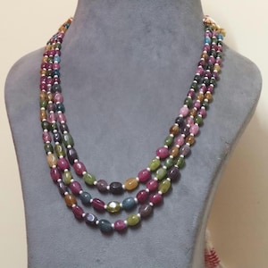 Op de afbeelding: Een ketting met meerdere strengen met ovaalvormige kralen in verschillende kleuren, waaronder roze, groen en geel. De kralen zijn geregen met kleine witte kralen. De ketting wordt getoond op een grijze mannequin.