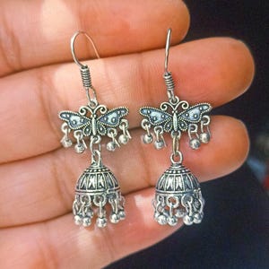 May include: Silver dangle earrings with a butterfly charm and a bell-shaped pendant. The earrings have a silver hook closure.