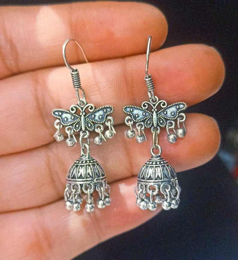 May include: Silver dangle earrings with a butterfly charm and a bell-shaped pendant. The earrings have a silver hook closure.