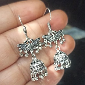 May include: Silver dangle earrings with a butterfly charm and a bell-shaped pendant. The earrings have a silver hook closure.