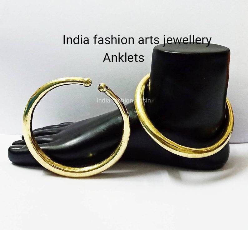 May include: Two gold-colored metal anklets on a black mannequin foot. The anklets are simple, round, and smooth. The text "India fashion arts jewellery Anklets" is visible above the foot.