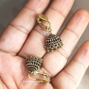 Könnte beinhalten: Ein Paar goldfarbene Ohrclips mit einem detaillierten, verzierten Design. Die Ohrringe haben einen glockenförmigen Anhänger mit einer strukturierten Oberfläche.