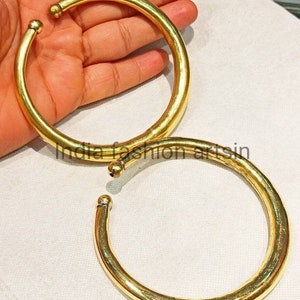 May include: Two gold-colored metal bangles, one slightly larger than the other, are shown on a white surface. The bangles are smooth and round.