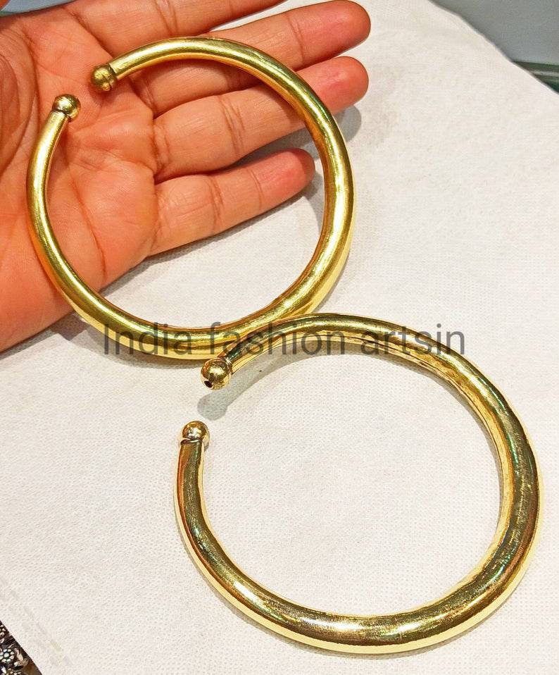 May include: Two gold-colored metal bangles, one slightly larger than the other, are shown on a white surface. The bangles are smooth and round.