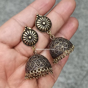 May include: A pair of gold-toned earrings with a round, ornate design and a dangling bell-shaped charm. The earrings are made of metal and have a traditional Indian style.