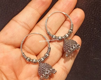 May include: A pair of silver hoop earrings with dangling bell-shaped charms. The hoops are adorned with small beads and the charms have intricate detailing. The earrings are displayed against a dark background.