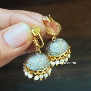 Könnte beinhalten: Goldfarbene Clip-Ohrringe mit hellblauem, gemustertem Kuppeldesign. Die Ohrringe haben eine goldene Filigranbasis mit baumelnden weißen Perlen. Die Ohrringe werden in einer Hand gehalten.