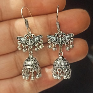 May include: Silver dangle earrings with a butterfly charm and a bell-shaped pendant. The earrings have a silver finish and are decorated with intricate details.