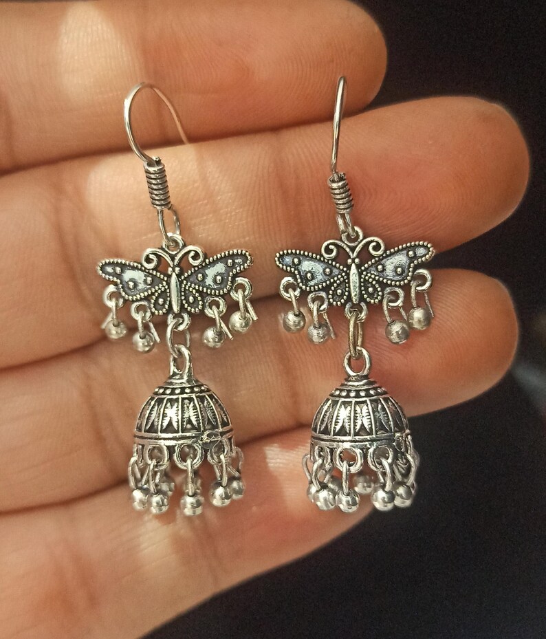 May include: Silver dangle earrings with a butterfly charm and a bell-shaped pendant. The earrings have a silver finish and are decorated with intricate details.