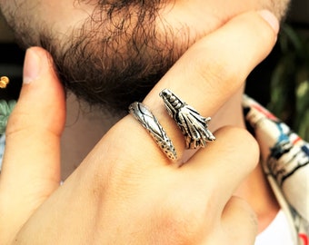 Op de afbeelding: Een zilverkleurige ring met een drakenontwerp. De ring heeft een band met textuur en een drakenkop aan de voorkant. De kop van de draak is gedetailleerd met schubben en een felle uitdrukking. De ring wordt op een vinger gedragen.