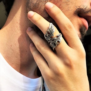 Puede incluir: Un anillo de plata con un diseño de dos plumas superpuestas. El anillo se lleva en el dedo índice de una mano.