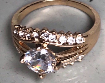 Op de afbeelding: Een gouden ring met een grote, heldere steen en kleinere, heldere stenen die in een band rond de steen zijn gezet.