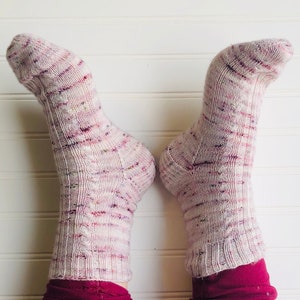 May include: A pair of pink and white patterned socks with a cable knit design. The socks are worn by a person wearing burgundy pants.