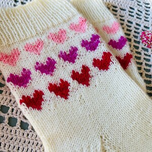 May include: A pair of white knitted socks with a pattern of pink, purple, and red hearts. The socks are on a crocheted surface.