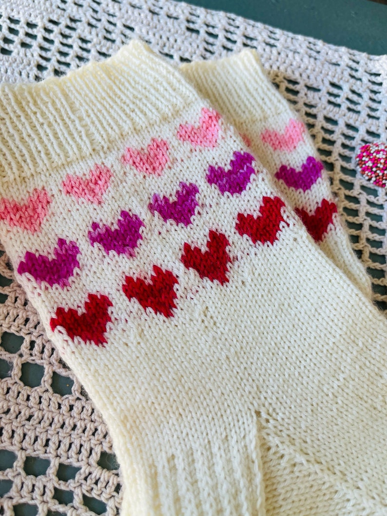 May include: A pair of white knitted socks with a pattern of pink, purple, and red hearts. The socks are on a crocheted surface.