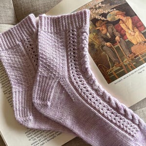 Peut inclure: Une paire de chaussettes tricotées lavande avec des motifs de câbles et de dentelle. Les chaussettes sont posées sur un livre ouvert avec une illustration. Les chaussettes sont en fil doux.
