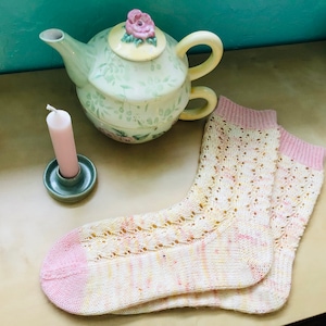 Pode incluir: Um par de meias de tricô rosa e amarelo com um padrão de renda. As meias estão sobre uma superfície marrom claro ao lado de um castiçal verde e uma chaleira floral.