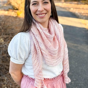 May include: A light pink knitted shawl with a delicate lace pattern is draped over a white top and a pink skirt. The shawl has a scalloped edge and is worn by a person with long brown hair.