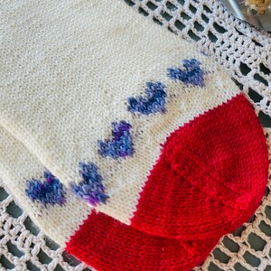 May include: A pair of white knitted socks with a red heel and toe. The socks have a pattern of blue hearts on the foot.