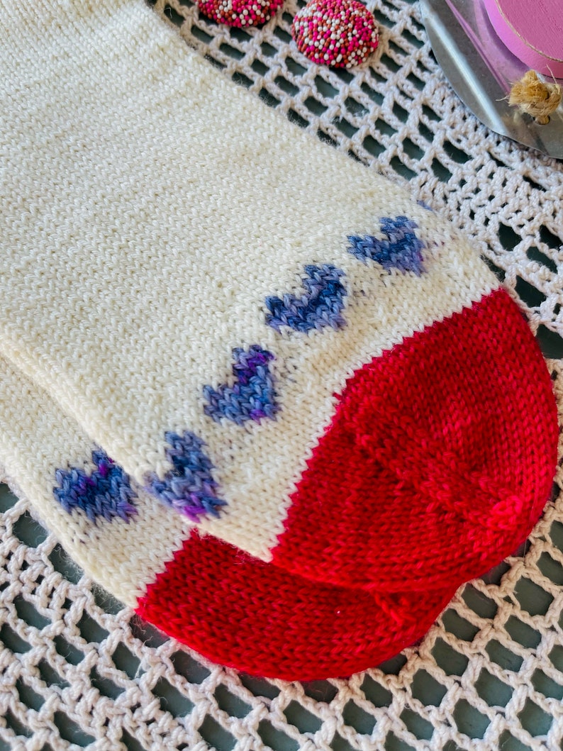 May include: A pair of white knitted socks with a red heel and toe. The socks have a pattern of blue hearts on the foot.