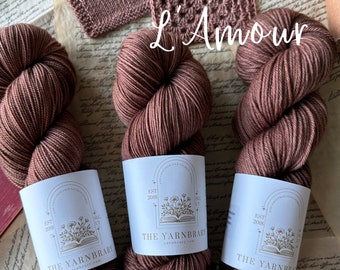 May include: Three skeins of L'Amour yarn in a deep rose color. Each skein is wrapped around a white label with the text "The Yarnbrary" and a floral design.